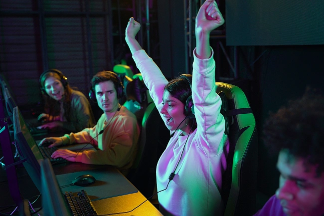 Een jonge vrouwelijke gamer viert enthousiast met haar handen in de lucht in een internetcafé, omringd door andere spelers met headsets.